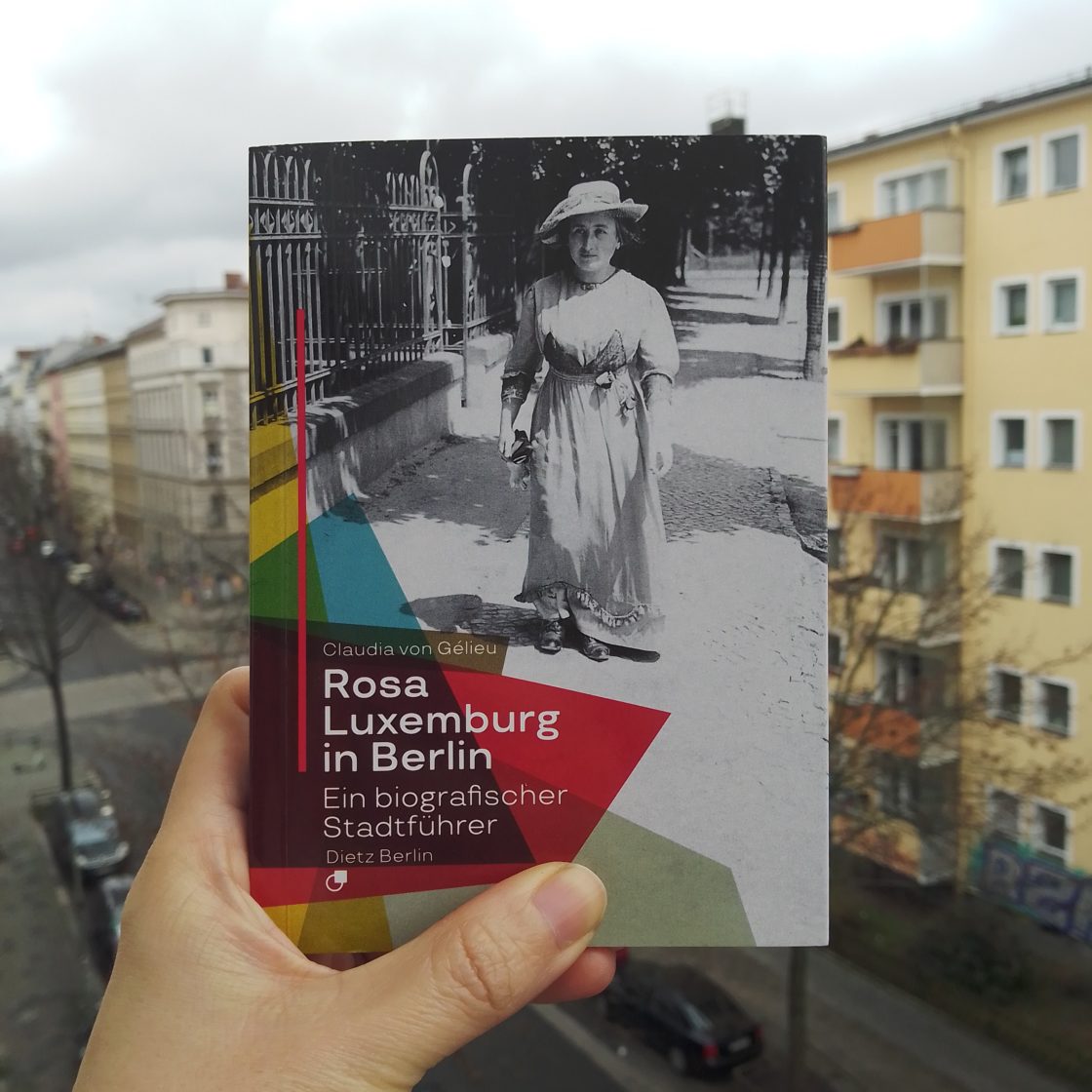 book, Rosa Luxemburg in Berlin hold with a left hand, in front of pale yellow building with many windows. On the cover, Rosa Luxemburg in summer dress and hat walking on a street