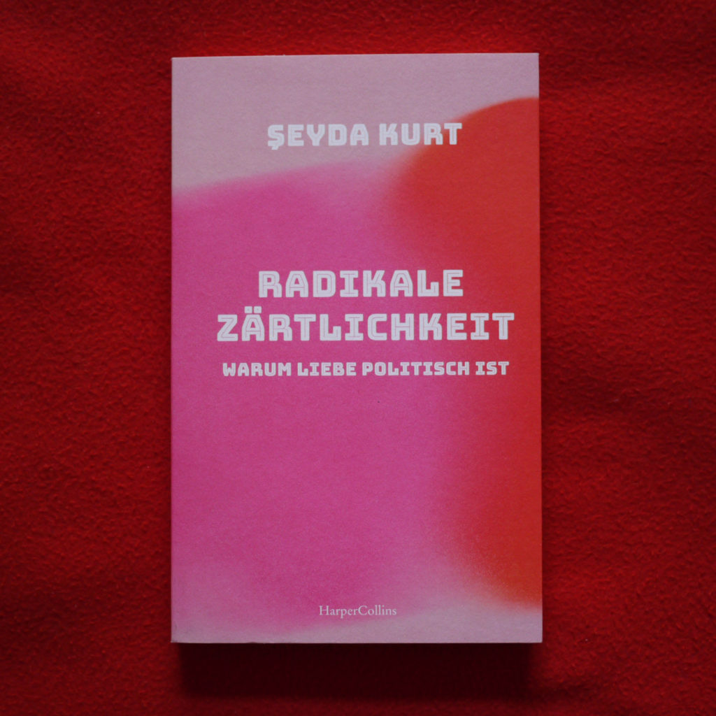 Auf dem roten weichen Stoff liegt das Buch "Radikale Zärtlichkeit" von Şeyda Kurt. Das buch hat hell rosa cover, etewas dunklere pinke Kreis vom links, rote Kreis vom rechts, beide etwas verschwommen und verschmelzen ineinander