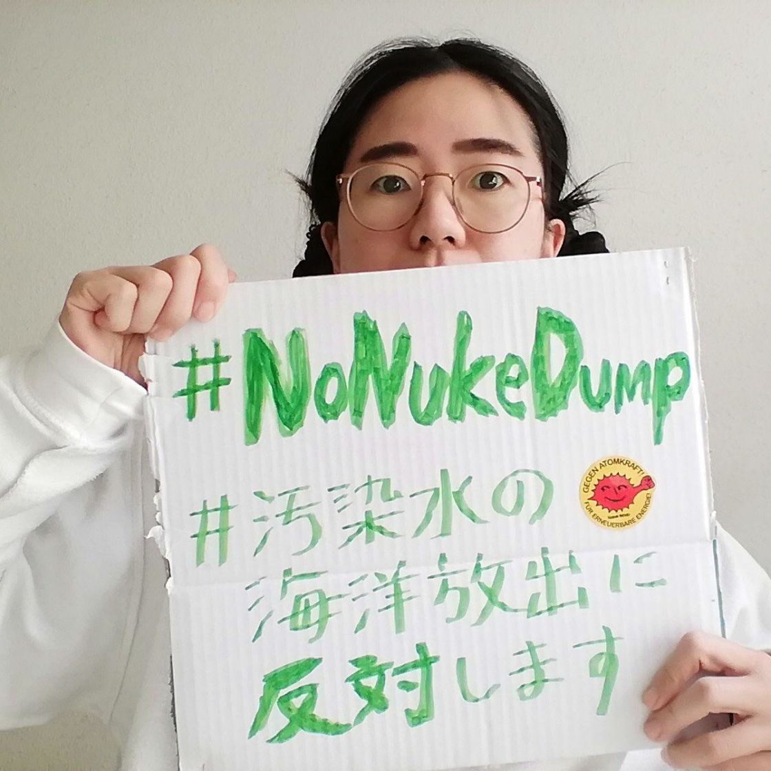 Aiko hält ein Schild mit den grünen Schriften #NoNukeDump, #汚染水の海洋放出に反対します mit einem gelben Aufkleber "Gegen Atomkraft" Für erneuerbare Energie" in der Mitte rote zackig rundes Gesicht mit gehobenem Faust