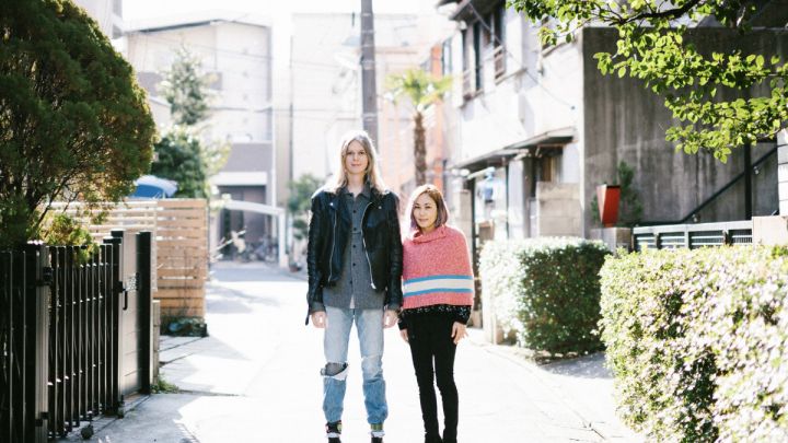 Elin McCready and Midori Morita standing in a surbian street in Japan, full of lights. Elin wears black leather jacket and jeans. She is tall and has silver long hair. Midori has pink knit poncho with light blue and white stripe below with black skinny pants. She has blond shoulder length hair, is high as shoulder of Elin. by Yasuyo Tadokoro