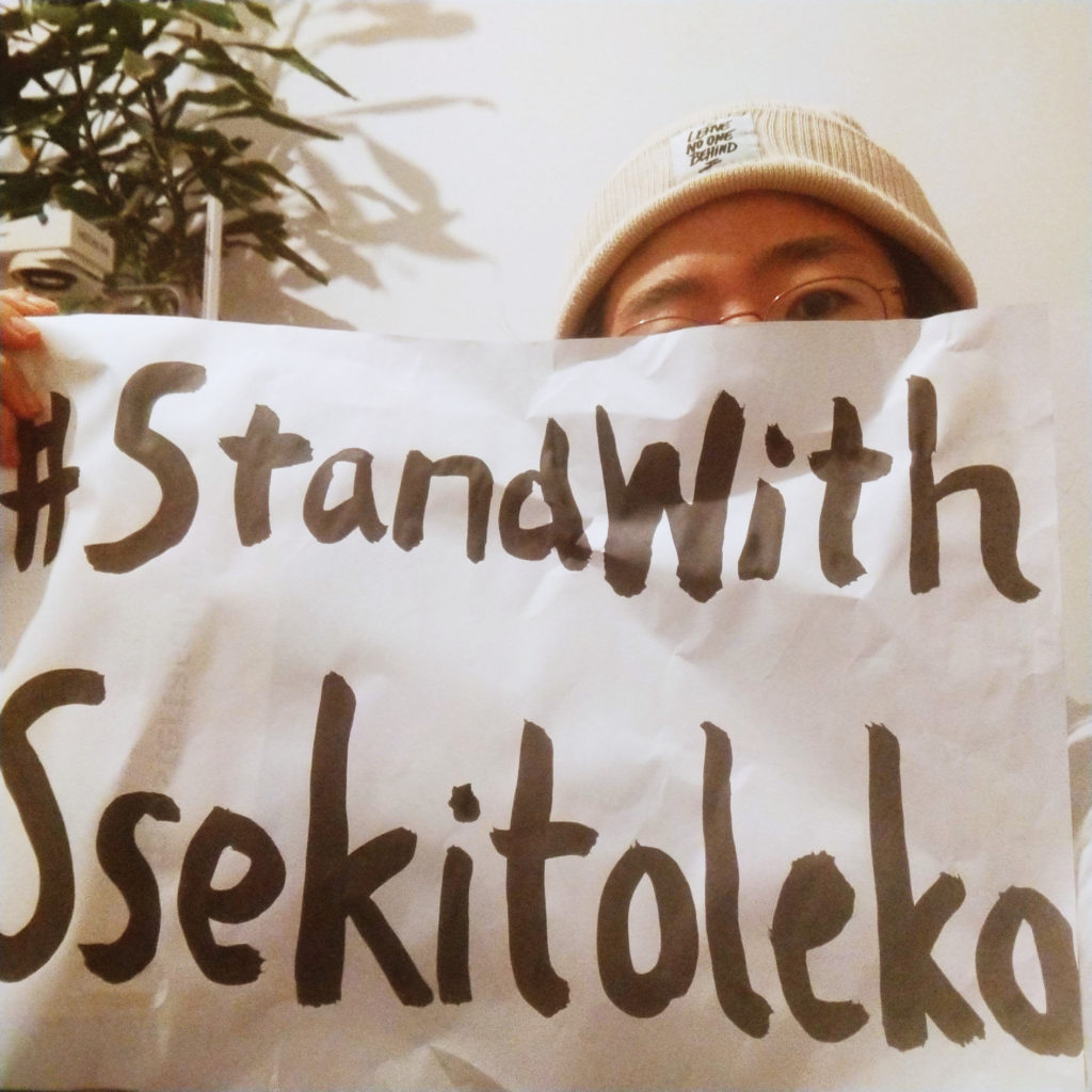 A person with a beanie with a patch "leave no one behind" holding a paper with #StandWithSsekitoleko, written with japanese caligraphy.