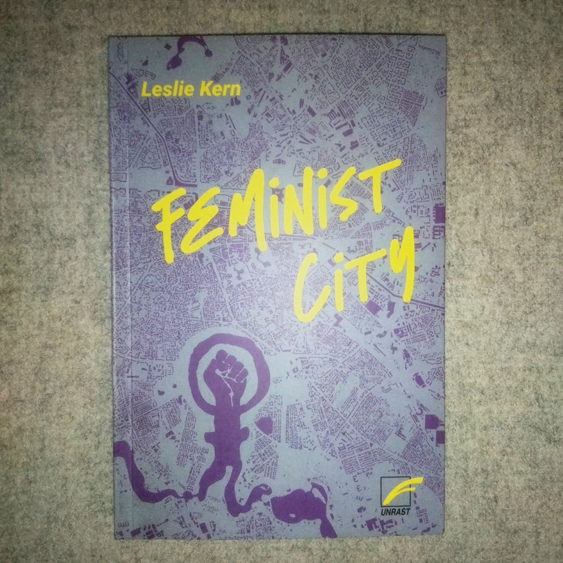 cover von dem Buch "Feminist City" von Leslie Kern. Auf hellgrauem Hintergrund in lila Stadtplan, Faust und Frau Zeichen, Titel in neon gelb, unten Rechts ein Logo von UNRAST