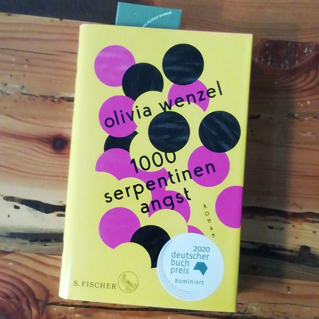 Das Buch, 1000 Serpentinen Angst von Olivia Wenzel mit dem gelbem Cover mit pink und schwarze Kreise auf einem Holztisch